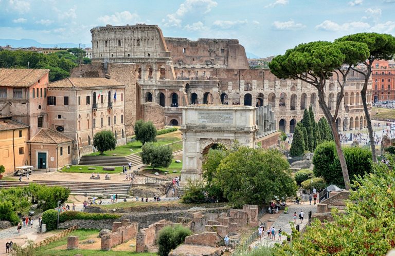 Foto de Roma. Ruinas, edificios ao fundo e pessoas caminhando na praça.