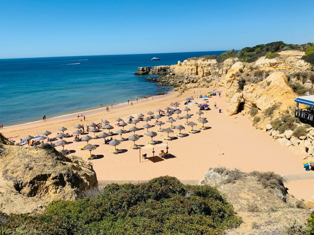 Algarve. Foto tirada de cima com praia embaixo, guarda-sóis e algumas pessoas.