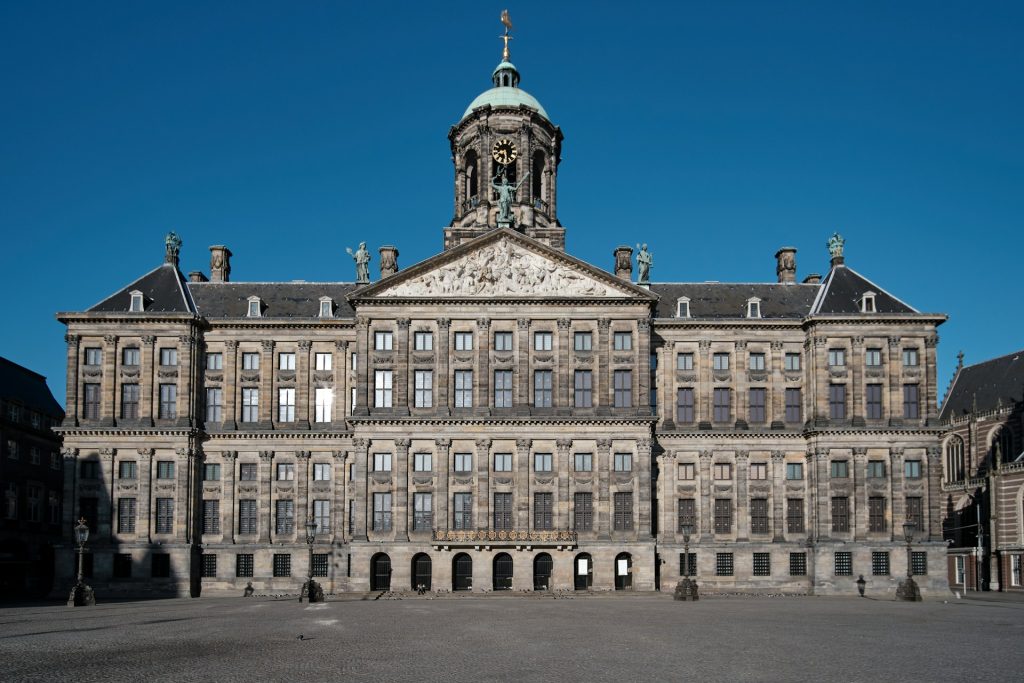 Palacio Real Amsterda. Praça com grande edifício marrom com muitas janelas. 