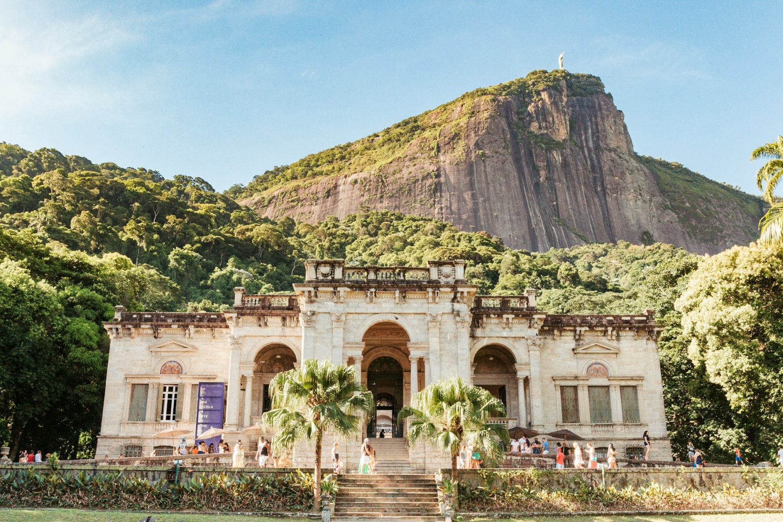 Parque Lage. Edificio de concreto branco com montanha ao fundo e pessoas caminhando na frente