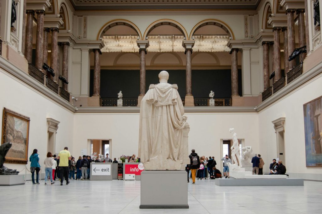 Museu Bruxelas. Foto dentro de uma sala com uma grande estatua de concreto branco e pessoas caminhando dentro do museu.