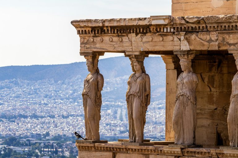 Athenas. Foto de um grupo de estatuas na leteral de um edificio e a cidade de athenas ao fundo.