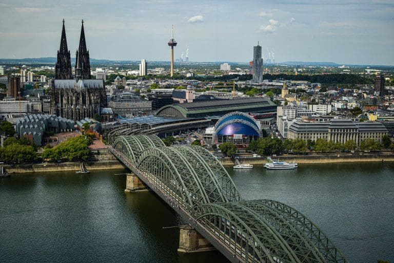 Colonia. Foto tirada de cima com uma ponte de metal sobre corpo de agua e cidade na margem.