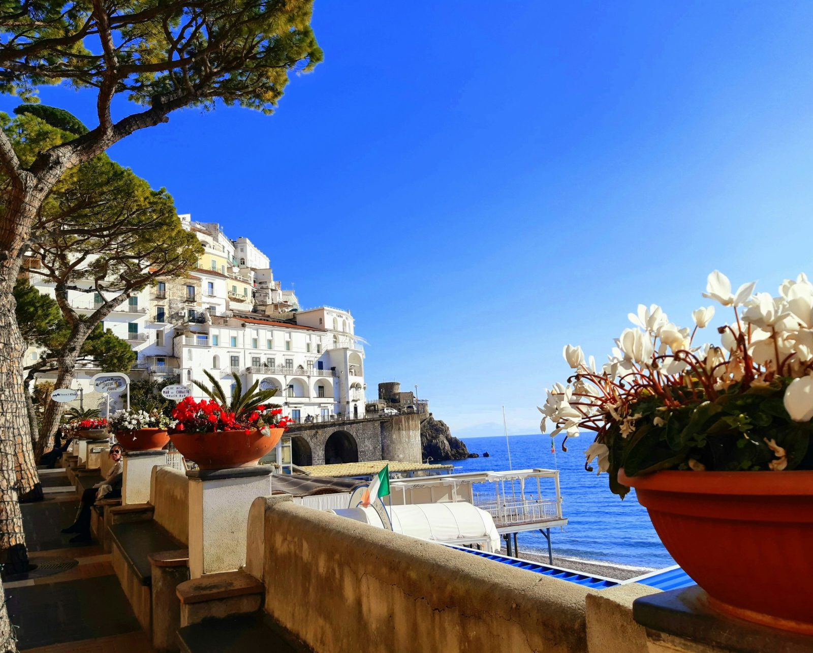 Foto Amalfi. Vista com vaso de flores, edifício branco e mar azul ao fundo.